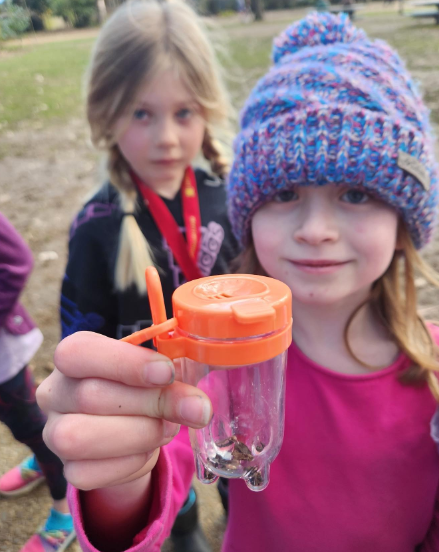 An elementary school student holds up a jar containing an insect they caught.